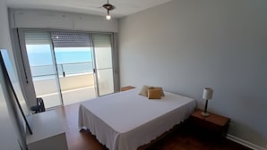 A bedroom with a bed, a nightstand, a lamp, and a view of the sea through a sliding door. - SHB - Facing the Sea at Pitangueiras Beach (Guarujá)