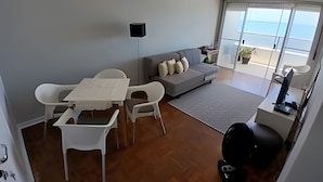 A modern living room with a grey sofa, white chairs, a small table, and a view of the sea through large glass doors. - SHB - Facing the Sea at Pitangueiras Beach (Guarujá)