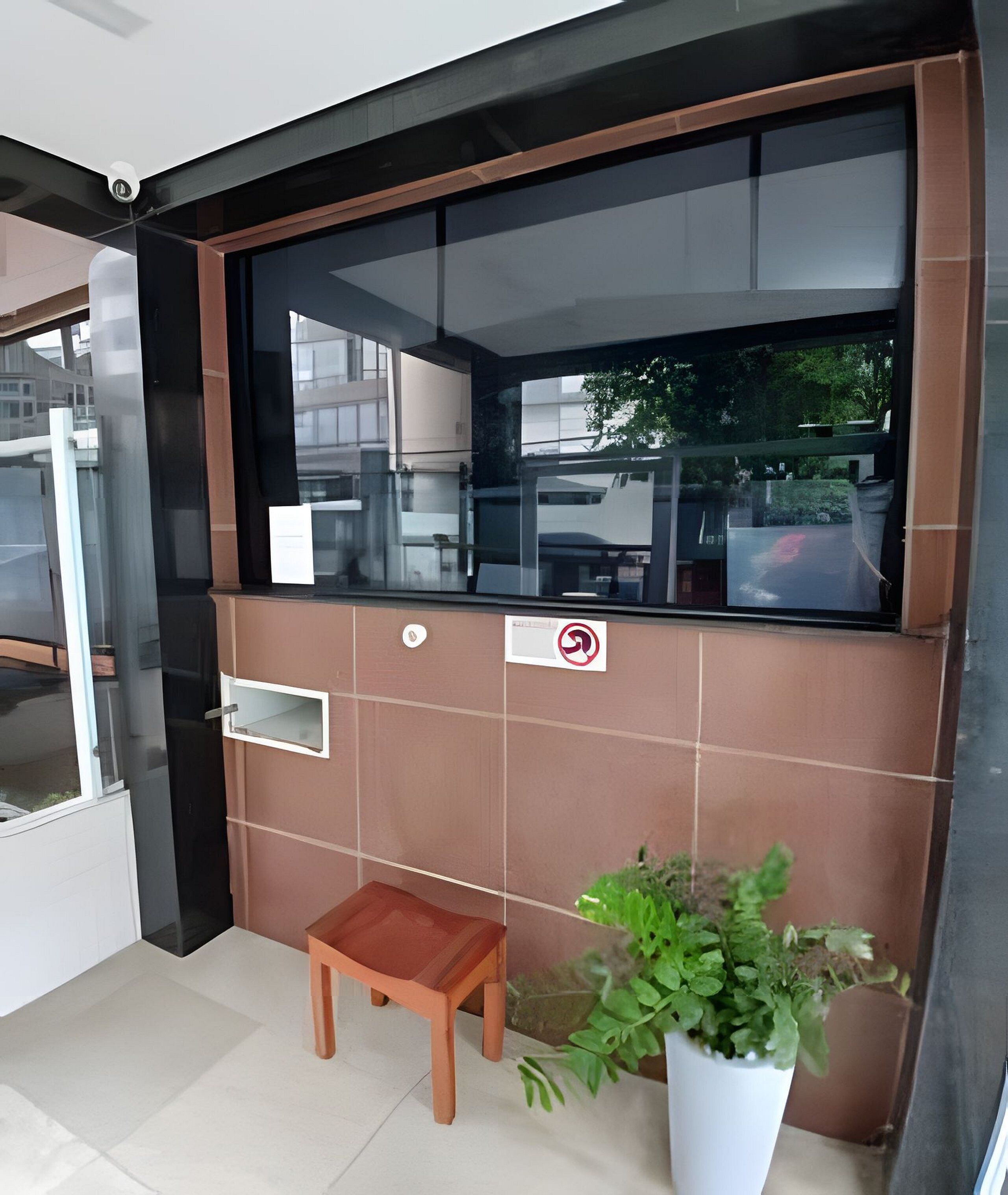 Un área de recepción con una ventana de vidrio, un taburete de madera, una planta en maceta y un letrero de prohibido fumar.