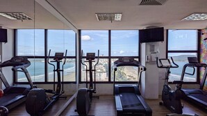Een sportschool met cardiotoestellen en een groot raam met uitzicht op een strand en stadsbeeld.