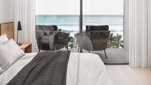 Quarto moderno com uma cama grande, mesa de cabeceira, luminária e duas cadeiras voltadas para uma varanda com vista para o oceano.