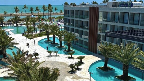 Resort com ampla área de piscina, palmeiras e um edifício com vários andares.