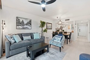 A modern living room with a grey sofa, a coffee table, and a dining area with a table and chairs. - H-346 -Third Floor - Sunset Views From The Lanai! (Naples)