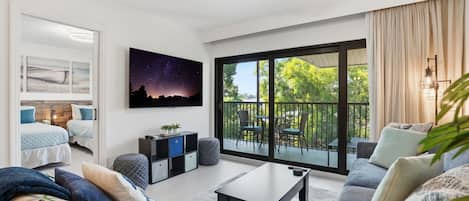 Una sala moderna con sofá, una mesa de centro y televisor montado en la pared. Se ve un balcón con mobiliario exterior a través de los amplios ventanales.