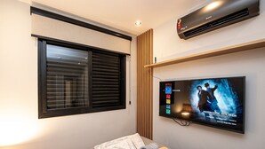 A hotel room with a bed, a TV displaying a show, a window with blinds, and an air conditioner. - SHB • Praiamar Shopping | Beach Just Steps Away (Santos)