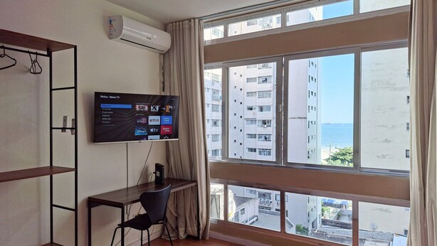 A hotel room with a large window, a flat-screen TV mounted on the wall, a desk with a chair, and a bed with a white duvet. - SHB - Maritime Comfort: Studio with Sea View (Santos)