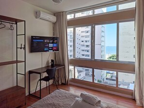 A hotel room with a large window, a flat-screen TV mounted on the wall, a desk with a chair, and a bed with a white duvet. - SHB - Maritime Comfort: Studio with Sea View (Santos)
