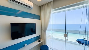 A modern room with a large TV mounted on a blue and white wall, a ceiling air conditioner, a glass door leading to a balcony with a view of the sea, and a blue chair on the balcony. - SHB - Wake Up to the Blue of the Sea at Your Window! (Guarujá)