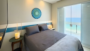 A bedroom with a bed, bedside tables, a wall decoration, and a view of the beach through a sliding door. - SHB - Wake Up to the Blue of the Sea at Your Window! (Guarujá)