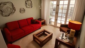 Un salon avec un canapé rouge, une table basse en bois et une lampe. On y trouve des œuvres d’art murales abstraites et une vue à travers une porte vitrée.