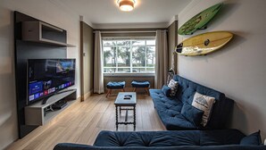 Una sala de estar con televisor, un sofá, una mesa de centro y dos sillas. Hay una ventana con cortinas y una tabla de surf en la pared.