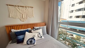 A bedroom with a wooden headboard, a macrame wall hanging, and a view of a building through a window. - SHB - The Best of Pitangueiras with Beach Service (Guarujá)