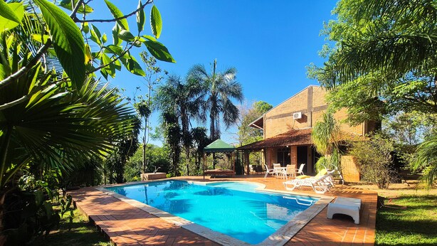 Une piscine rectangulaire bordée de carreaux, entourée d’une végétation luxuriante et d’une maison en briques.