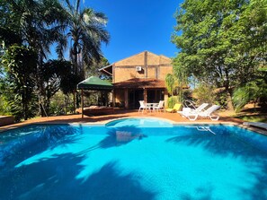 Une piscine avec des chaises longues, un espace de détente couvert et un bâtiment doté d’un toit en tuiles.