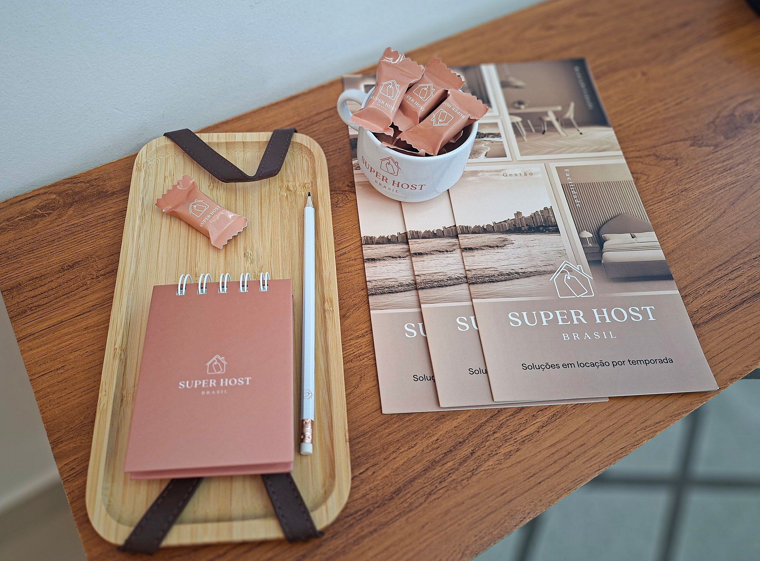 A wooden table with a notebook, a pen, a cup with chocolates, and brochures for Super Host Brasil.
