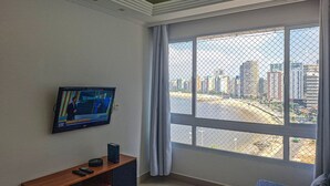 A room with a TV mounted on the wall, a wooden entertainment unit, a large window with a view of a cityscape, and a patterned rug.