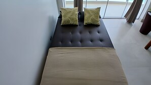 A bed with a dark headboard and two patterned pillows, a beige bedspread, and a view of the outdoors through large windows. - SHB - Amazing view | On the Sand | Pitangueiras (Guarujá)
