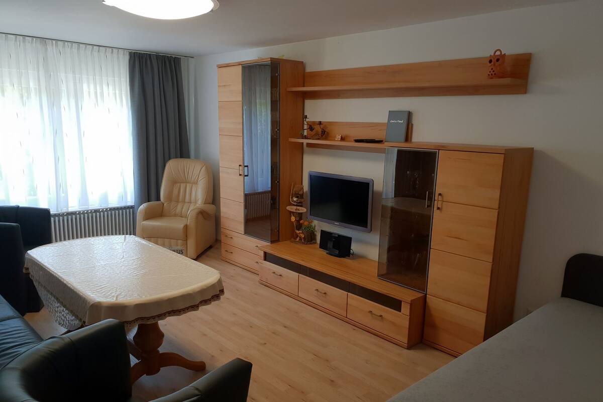 Ein Wohnzimmer mit einer Couch, einem Sessel, einem Fernseher, einer Holzkommode und einem Esstisch.