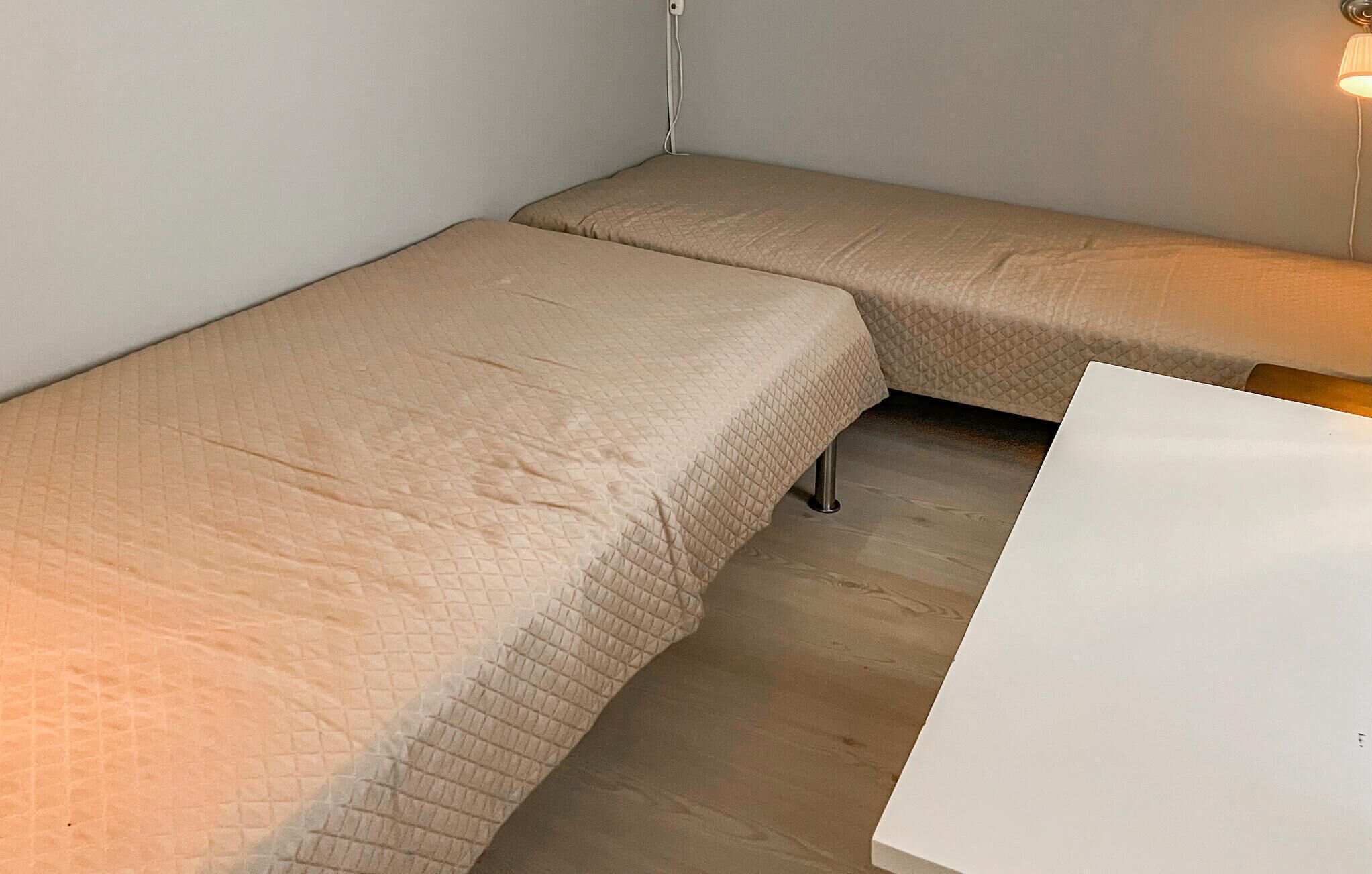 A small, compact room with two single beds, a white table, and a wall-mounted lamp.