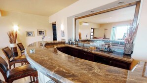 A modern kitchen with a curved granite countertop, a large mirror, and dark wooden chairs. - Golf & Beach Club Villa | Perfect for Families (Ciudad de los cabos)