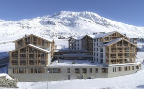 A multi-story lodge with a symmetrical design, featuring large windows and wooden balconies, set against a snowy mountain backdrop. - Ski-in/ski-out & foot of the slopes, fitness, swimming pool, sauna, hammam, terrace, tv, ski locker (Huez)