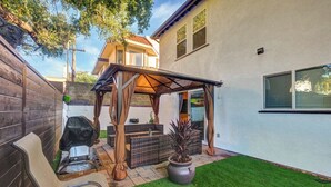 Un patio trasero con un porche de madera, mobiliario exterior, una parrilla y una planta en maceta.