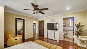Un dormitorio con cama, ventilador de techo, televisor y una cocina visible a través de una puerta abierta.