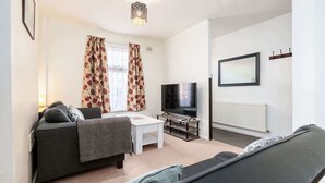 Ein Wohnzimmer mit einer Couch, einem Fernseher, einem weißen Tisch und einem Fenster mit Vorhängen.