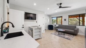 Eine moderne Küche mit weißen Schränken, einem schwarzen Wasserhahn und einem integrierten Fernseher. Im Wohnbereich befindet sich ein graues Sofa und ein kleiner Tisch, darüber ein Deckenventilator.
