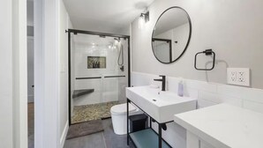 Ein modernes Badezimmer mit Duschkabine aus Glas, einem weißen Waschbecken mit schwarzem Wasserhahn und einem runden Spiegel an der Wand.