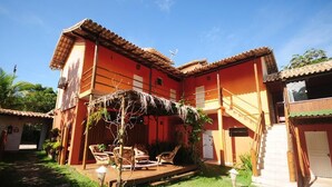 A two-story building with a red facade, a covered patio with chairs, and a staircase leading to the upper floor. - Mandacaru Pousada (Porto Seguro)