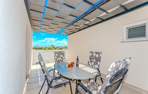 Eine überdachte Terrasse im Freien mit Tisch und Stühlen, einer Weinflasche und Blick auf Himmel und Gebäude im Hintergrund.