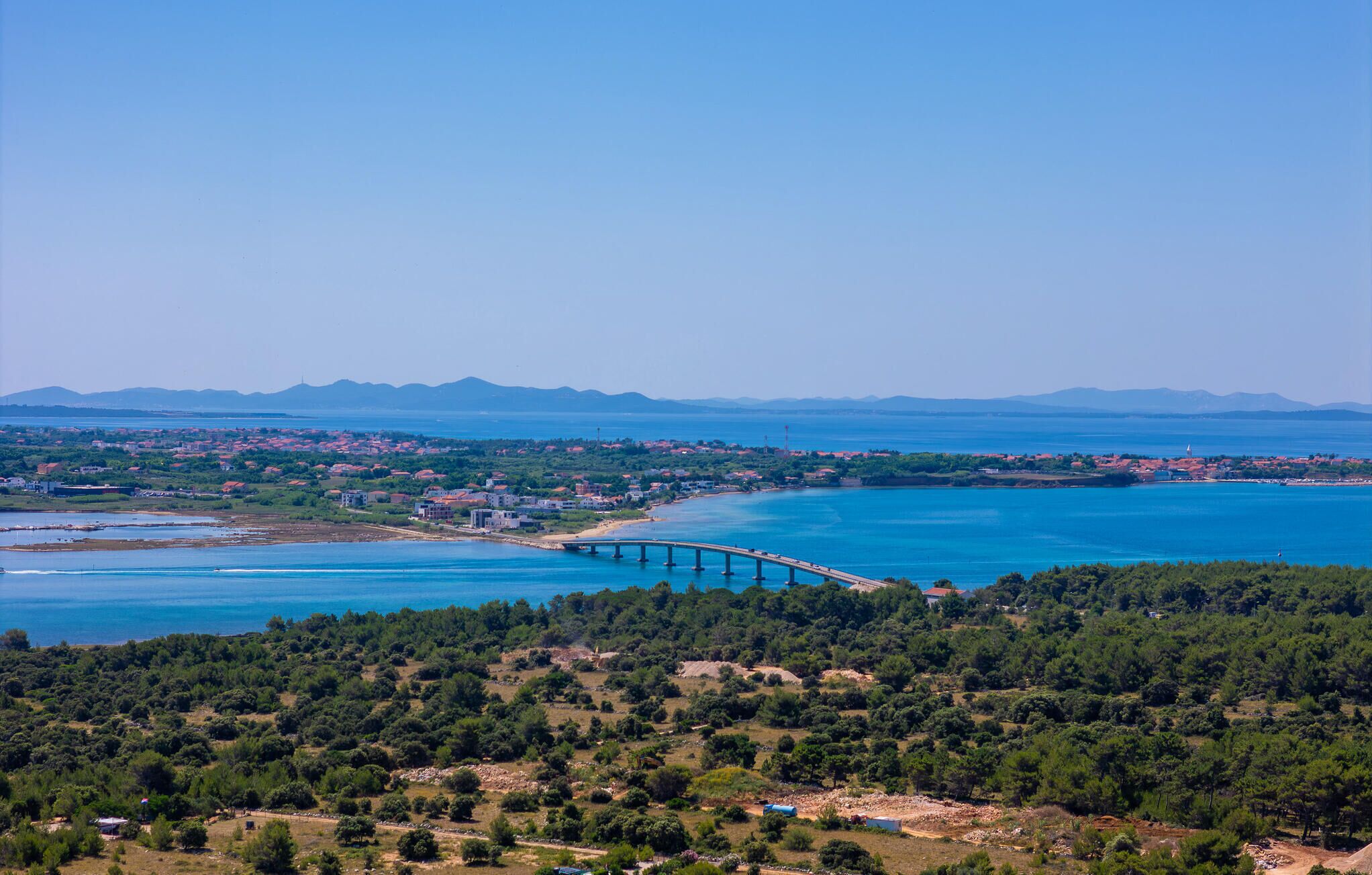 A coastal landscape with a bridge, greenery, and distant mountains.