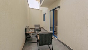 Un estrecho pasillo exterior con una zona de estar que incluye un sofá, sillas, una mesita y una ventana con persianas.