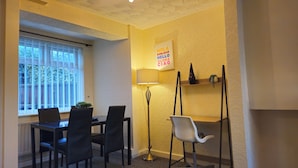 Ein Zimmer mit Esstisch, Stühlen, einer Stehlampe, einem Regal mit Dekoration und einem Fenster mit Jalousien.