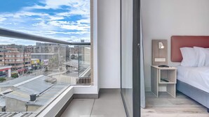 A modern hotel room with a large window offering a city view, a neatly made bed, a bedside table with a lamp, and a small shelf. - w* | Warm Loft w/ Balcony in Santa Barbara (Bogotá)