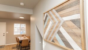 Ein Essbereich mit Wandkunst aus Holz im Fischgrätmuster, einem Esstisch mit Stühlen und einem Fenster mit Jalousien.