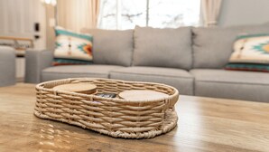Ein Korbkörbchen mit Holzdeckel auf einem Holztisch in einem Wohnzimmer mit grauem Sofa und gemusterten Kissen.