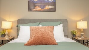 Een net opgemaakt bed met een groene plaid, twee kussens en een bruine deken. Twee nachtkastjes met lampen en kleine potplantjes staan ernaast. Boven het bed hangt een lijst met een berglandschap.