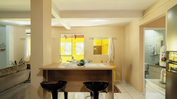 A compact kitchen with a central island, a dining area with two stools, a window with yellow curtains, and a bathroom visible to the right. - Apt w/ 2 Bedrooms and Barbecue Grill Fdc0005 (Florianópolis)
