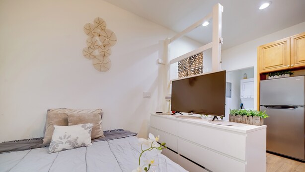 Une chambre moderne avec un lit, une table de chevet ornée d’un vase de fleurs, une télévision et un coin cuisine équipé d’un réfrigérateur et de placards.