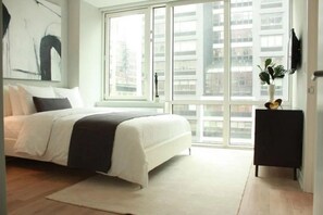 A modern bedroom with a large window, a bed, a black nightstand, and a painting on the wall. - Bright 1BR Chelsea Condo with Fitness Center (New York)