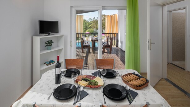 Un comedor con una mesa preparada para cuatro, un televisor en un estante y vistas a una terraza exterior con piscina y sillas.