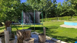 Un parque infantil con un trampolín y zona cubierta por red, rodeado de árboles y una piscina pequeña.