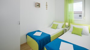 A hotel room with two single beds, each with a blue bedspread and green pillows. There is a small wooden shelf with a plant on the wall, and a window with green curtains. - Amazing home in Tinjan (Tinjan)
