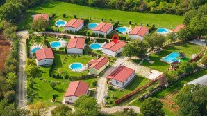 Oversigtsbillede af et boligområde med flere huse, alle med røde tage, omgivet af grønne græsplæner og træer. Flere swimmingpools er synlige, hvilket indikerer et fællesskab med fælles faciliteter.