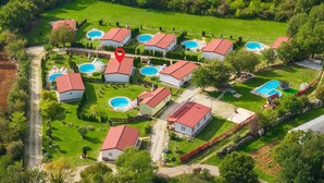 Vista aérea de um bairro residencial com várias casas, cada uma com telhado vermelho, cercadas por gramados verdes e árvores. Vários piscinas visíveis indicam uma comunidade com comodidades compartilhadas.
