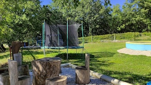 Legeplads med trampolin og netområde, omgivet af træer og en lille pool.