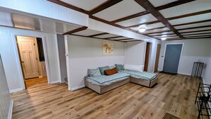 A room with a wooden floor, a sectional sofa with cushions, a door, and a ceiling with exposed beams. - 3 Bedroom house w/ lake access, hot tub and pet friendly! (Grawn)