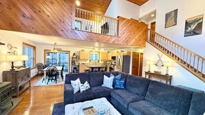 A spacious living area with a large gray sectional sofa, a round coffee table, and a staircase leading to an upper level. - Winnipesaukee Retreat | Hot Tub | Suissevale Beach (Moultonborough)